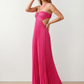 Woman wearing the Estella strapless pleated maxi dress featuring a ruched empire bodice, keyhole hardware detail, side cutout, open back with tie closure, and a flowing accordion-pleated floor-length skirt.