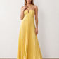 Woman wearing the Estella strapless pleated maxi dress featuring a ruched empire bodice, keyhole hardware detail, side cutout, open back with tie closure, and a flowing accordion-pleated floor-length skirt.