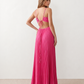 Woman wearing the Estella strapless pleated maxi dress featuring a ruched empire bodice, keyhole hardware detail, side cutout, open back with tie closure, and a flowing accordion-pleated floor-length skirt.