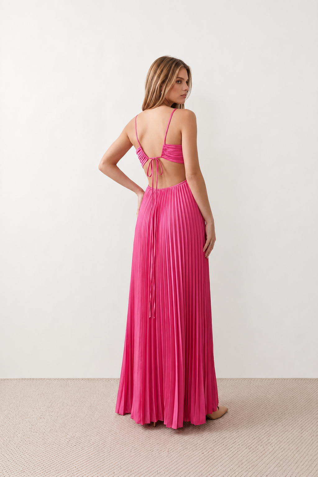 Woman wearing the Estella strapless pleated maxi dress featuring a ruched empire bodice, keyhole hardware detail, side cutout, open back with tie closure, and a flowing accordion-pleated floor-length skirt.