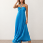 Woman wearing the Estella strapless pleated maxi dress featuring a ruched empire bodice, keyhole hardware detail, side cutout, open back with tie closure, and a flowing accordion-pleated floor-length skirt.