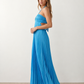 Woman wearing the Estella strapless pleated maxi dress featuring a ruched empire bodice, keyhole hardware detail, side cutout, open back with tie closure, and a flowing accordion-pleated floor-length skirt.