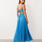 Woman wearing the Estella strapless pleated maxi dress featuring a ruched empire bodice, keyhole hardware detail, side cutout, open back with tie closure, and a flowing accordion-pleated floor-length skirt.