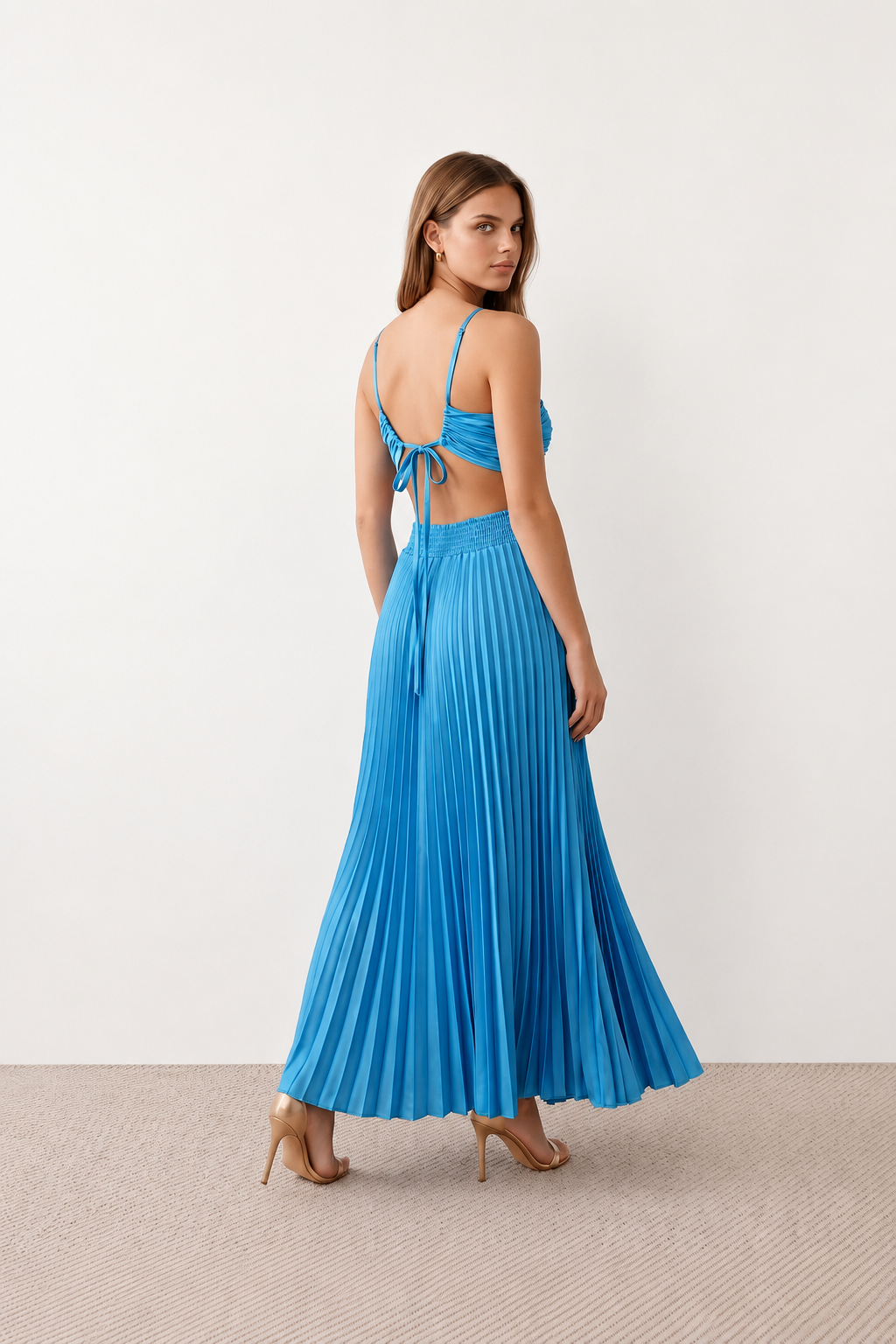 Woman wearing the Estella strapless pleated maxi dress featuring a ruched empire bodice, keyhole hardware detail, side cutout, open back with tie closure, and a flowing accordion-pleated floor-length skirt.