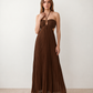 Woman wearing the Estella strapless pleated maxi dress featuring a ruched empire bodice, keyhole hardware detail, side cutout, open back with tie closure, and a flowing accordion-pleated floor-length skirt.