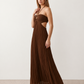 Woman wearing the Estella strapless pleated maxi dress featuring a ruched empire bodice, keyhole hardware detail, side cutout, open back with tie closure, and a flowing accordion-pleated floor-length skirt.