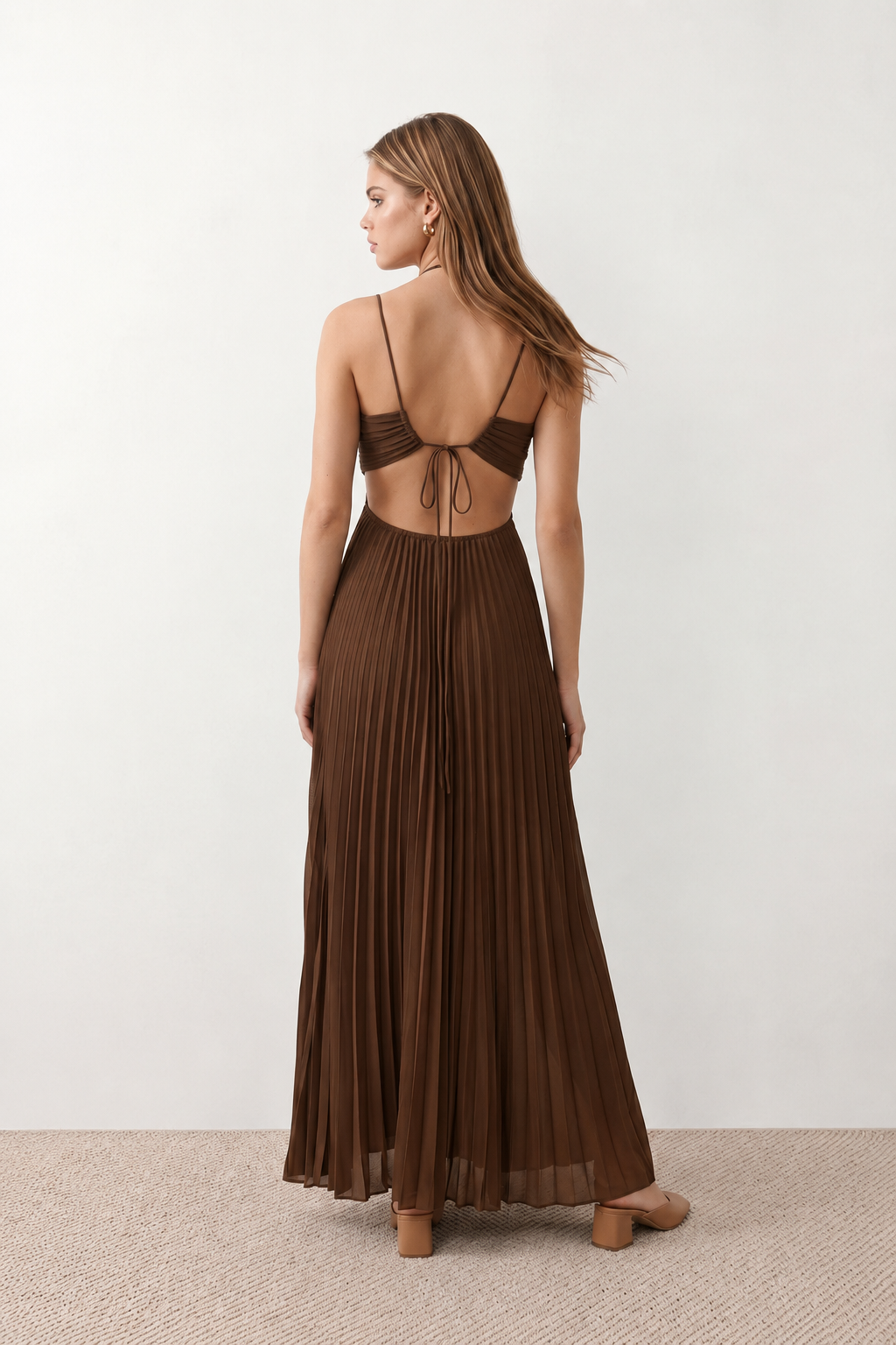 Woman wearing the Estella strapless pleated maxi dress featuring a ruched empire bodice, keyhole hardware detail, side cutout, open back with tie closure, and a flowing accordion-pleated floor-length skirt.