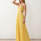 Woman wearing the Estella strapless pleated maxi dress featuring a ruched empire bodice, keyhole hardware detail, side cutout, open back with tie closure, and a flowing accordion-pleated floor-length skirt.