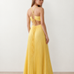 Woman wearing the Estella strapless pleated maxi dress featuring a ruched empire bodice, keyhole hardware detail, side cutout, open back with tie closure, and a flowing accordion-pleated floor-length skirt.