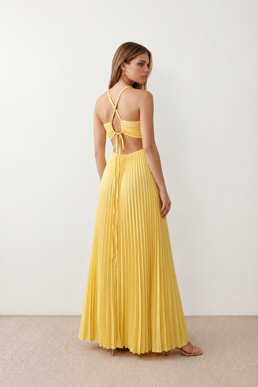 Woman wearing the Estella strapless pleated maxi dress featuring a ruched empire bodice, keyhole hardware detail, side cutout, open back with tie closure, and a flowing accordion-pleated floor-length skirt.