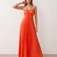 Woman wearing the Estella strapless pleated maxi dress featuring a ruched empire bodice, keyhole hardware detail, side cutout, open back with tie closure, and a flowing accordion-pleated floor-length skirt.