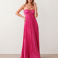 Woman wearing the Estella strapless pleated maxi dress featuring a ruched empire bodice, keyhole hardware detail, side cutout, open back with tie closure, and a flowing accordion-pleated floor-length skirt.