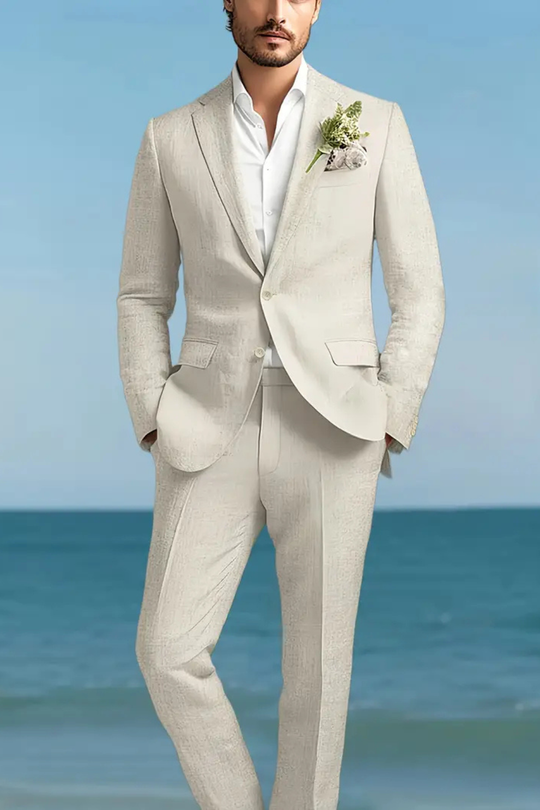 Man in a light gray suit standing on a beach with clear blue sky.