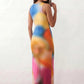 Woman wearing the Calia abstract print side slit maxi dress in a vibrant multicolour tie-dye design, featuring a sleeveless round neckline, body-skimming column silhouette, thigh-high side slit, and full-length flowing skirt, ideal for summer holidays, weddings, and occasion wear.