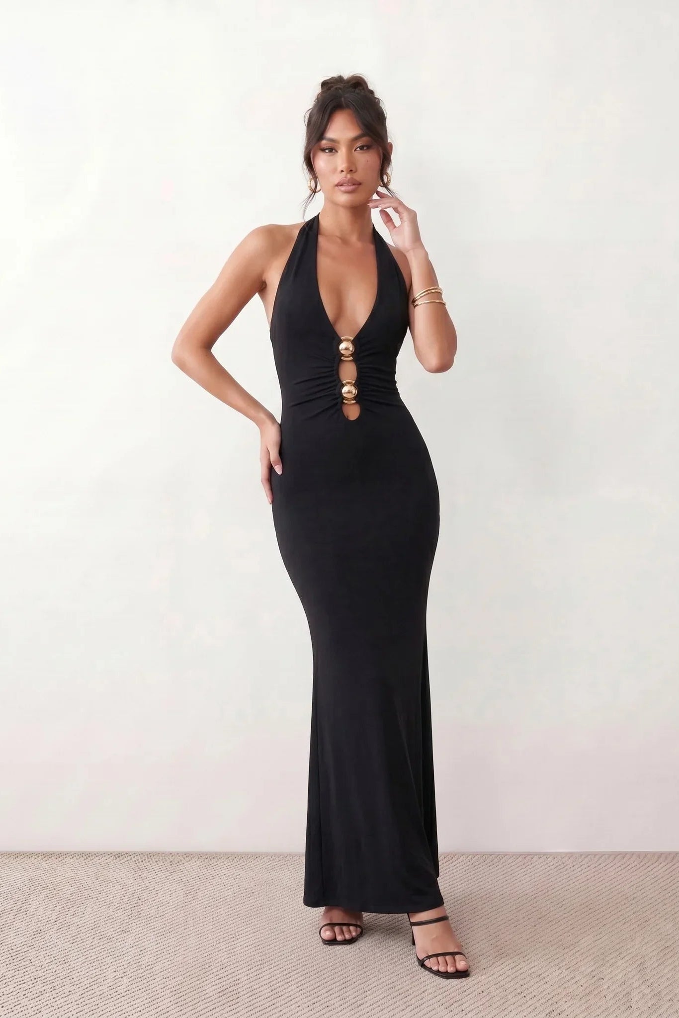Woman wearing the Ariadna Mermaid Halter Maxi Dress featuring a deep plunge halter neckline, open back with adjustable tie fastening, gold-tone hardware at the ruched waist, subtle waist cut-out and a floor-length mermaid silhouette, ideal for weddings, black-tie events and formal evening occasions.