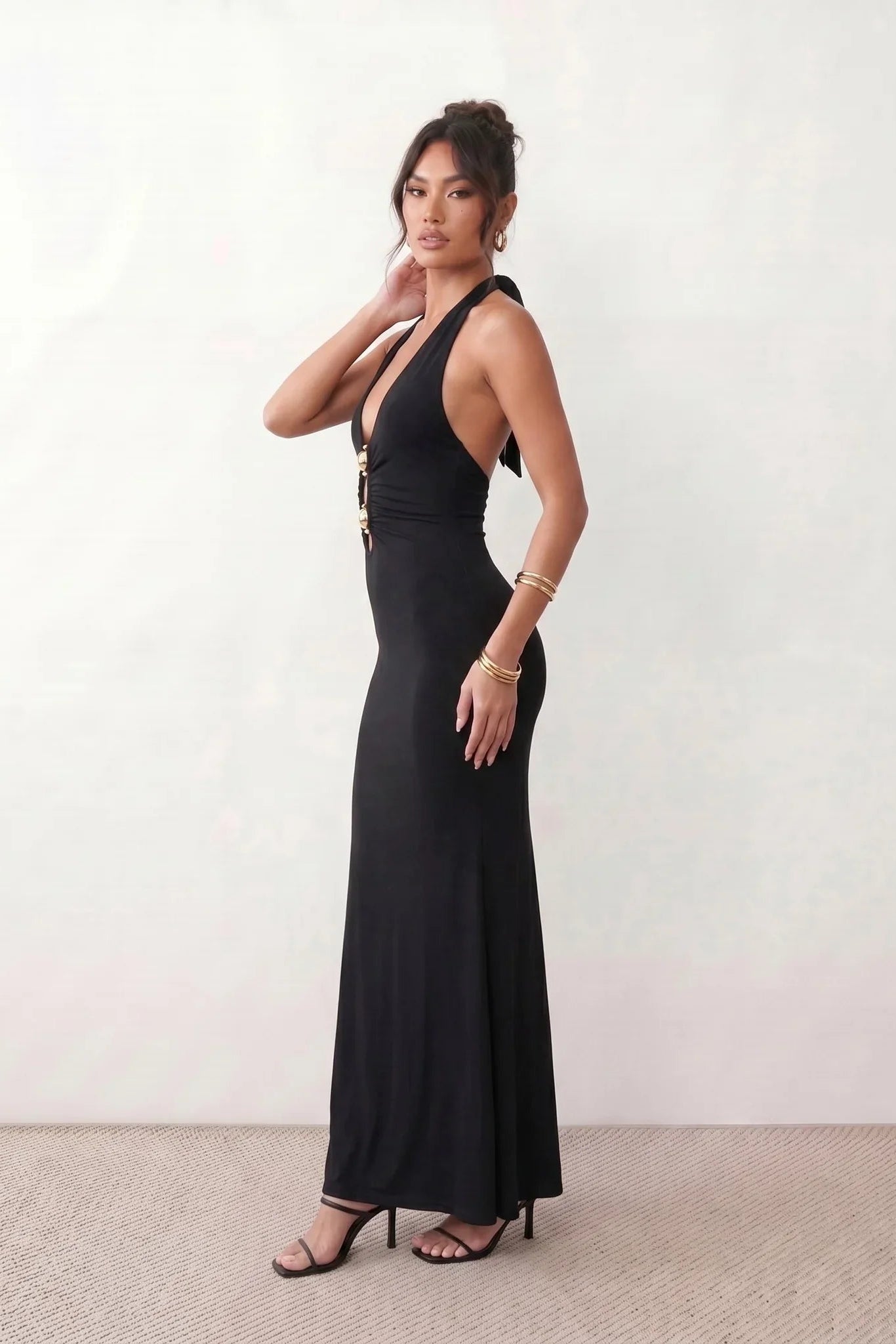 Woman wearing the Ariadna Mermaid Halter Maxi Dress featuring a deep plunge halter neckline, open back with adjustable tie fastening, gold-tone hardware at the ruched waist, subtle waist cut-out and a floor-length mermaid silhouette, ideal for weddings, black-tie events and formal evening occasions.