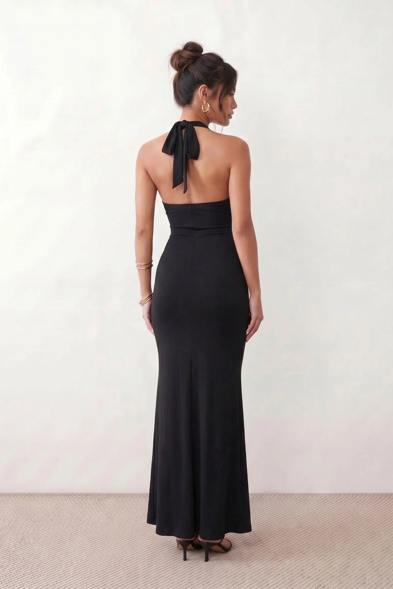 Woman wearing the Ariadna Mermaid Halter Maxi Dress featuring a deep plunge halter neckline, open back with adjustable tie fastening, gold-tone hardware at the ruched waist, subtle waist cut-out and a floor-length mermaid silhouette, ideal for weddings, black-tie events and formal evening occasions.