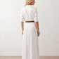 Woman wearing the Julieta Button Front Maxi Dress with a deep V-neck, shirt collar, button-front bodice, long sleeves, belted waist with a square-buckle belt, asymmetric pleated hip drape, and flowing maxi-length skirt.
