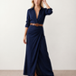 Woman wearing the Julieta Button Front Maxi Dress with a deep V-neck, shirt collar, button-front bodice, long sleeves, belted waist with a square-buckle belt, asymmetric pleated hip drape, and flowing maxi-length skirt.
