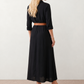 Woman wearing the Julieta Button Front Maxi Dress with a deep V-neck, shirt collar, button-front bodice, long sleeves, belted waist with a square-buckle belt, asymmetric pleated hip drape, and flowing maxi-length skirt.