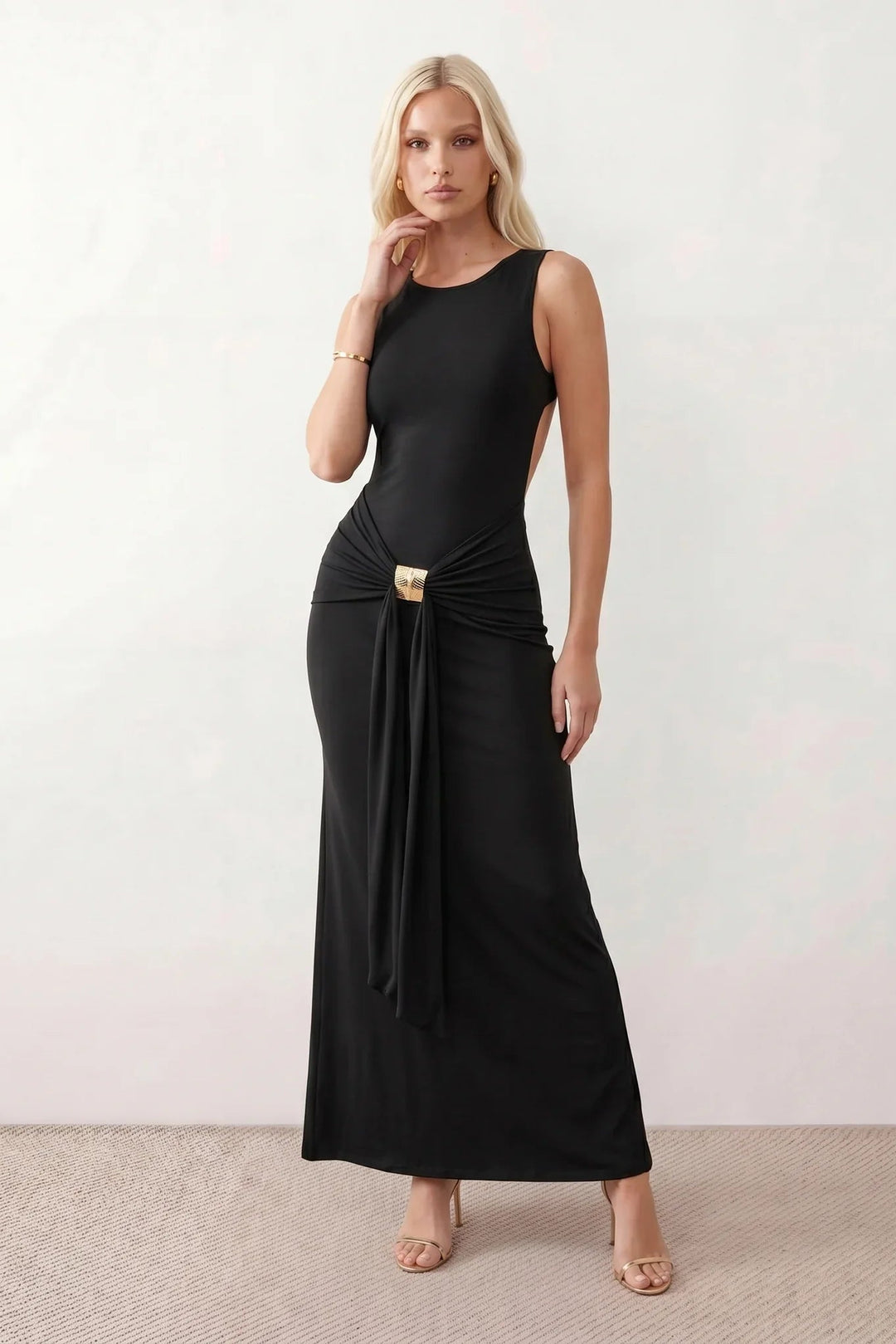 Woman wearing the Calista Open Back Fitted Maxi Dress in a fitted floor-length silhouette featuring a round neckline, sleeveless design, dramatic open back, ruched waist, gold-tone buckle detail at the hip, and a subtle mermaid hem, perfect for weddings, black-tie events and formal occasions.