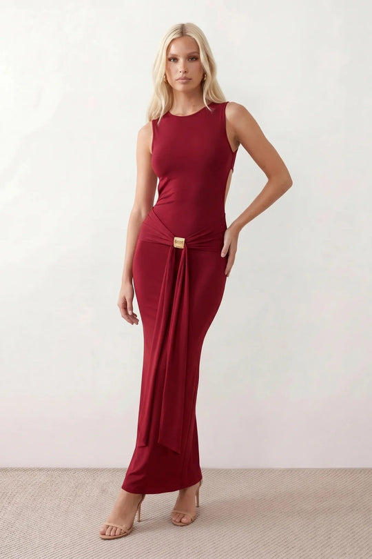 Woman wearing the Calista Open Back Fitted Maxi Dress in a fitted floor-length silhouette featuring a round neckline, sleeveless design, dramatic open back, ruched waist, gold-tone buckle detail at the hip, and a subtle mermaid hem, perfect for weddings, black-tie events and formal occasions.