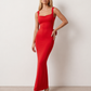 Woman wearing the Aline Cowl Neck Backless Maxi Dress in a floor-length fitted silhouette with a draped cowl neckline, elegant open back, oversized satin bow tie detail, sleeveless design, and smooth woven fabric, ideal for weddings, black-tie events, galas, and evening occasions.