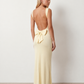 Woman wearing the Aline Cowl Neck Backless Maxi Dress in a floor-length fitted silhouette with a draped cowl neckline, elegant open back, oversized satin bow tie detail, sleeveless design, and smooth woven fabric, ideal for weddings, black-tie events, galas, and evening occasions.