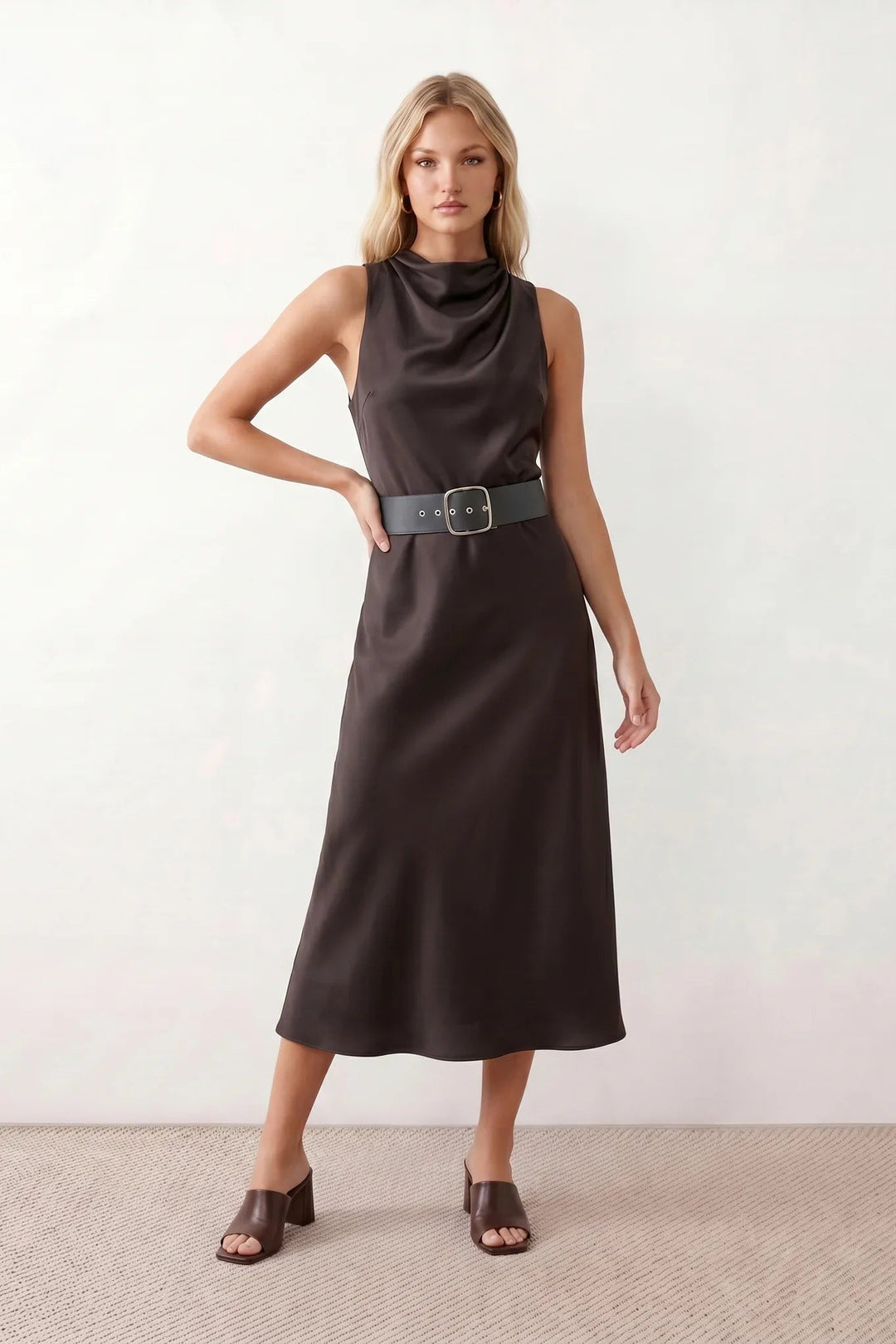 Woman wearing the glossy cowl neck belted midi dress featuring a draped neckline, sleeveless sheath silhouette, removable wide buckle belt, and elegant midi-length hem in a lustrous finish.