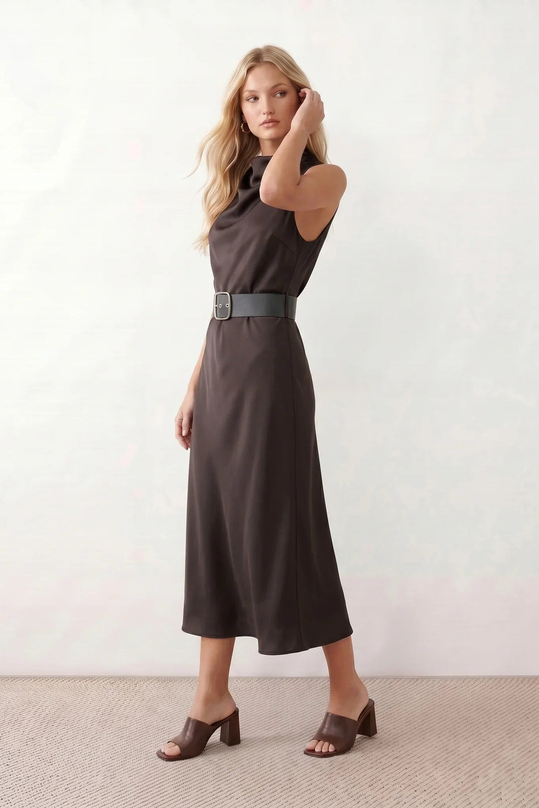 Woman wearing the glossy cowl neck belted midi dress featuring a draped neckline, sleeveless sheath silhouette, removable wide buckle belt, and elegant midi-length hem in a lustrous finish.