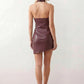 Woman wearing a glossy faux leather halter mini dress with a draped cowl neckline, fully backless design, ruched waist and hips, bodycon fit, and an asymmetric mini hem, styled for parties, cocktail events, and evening occasions.