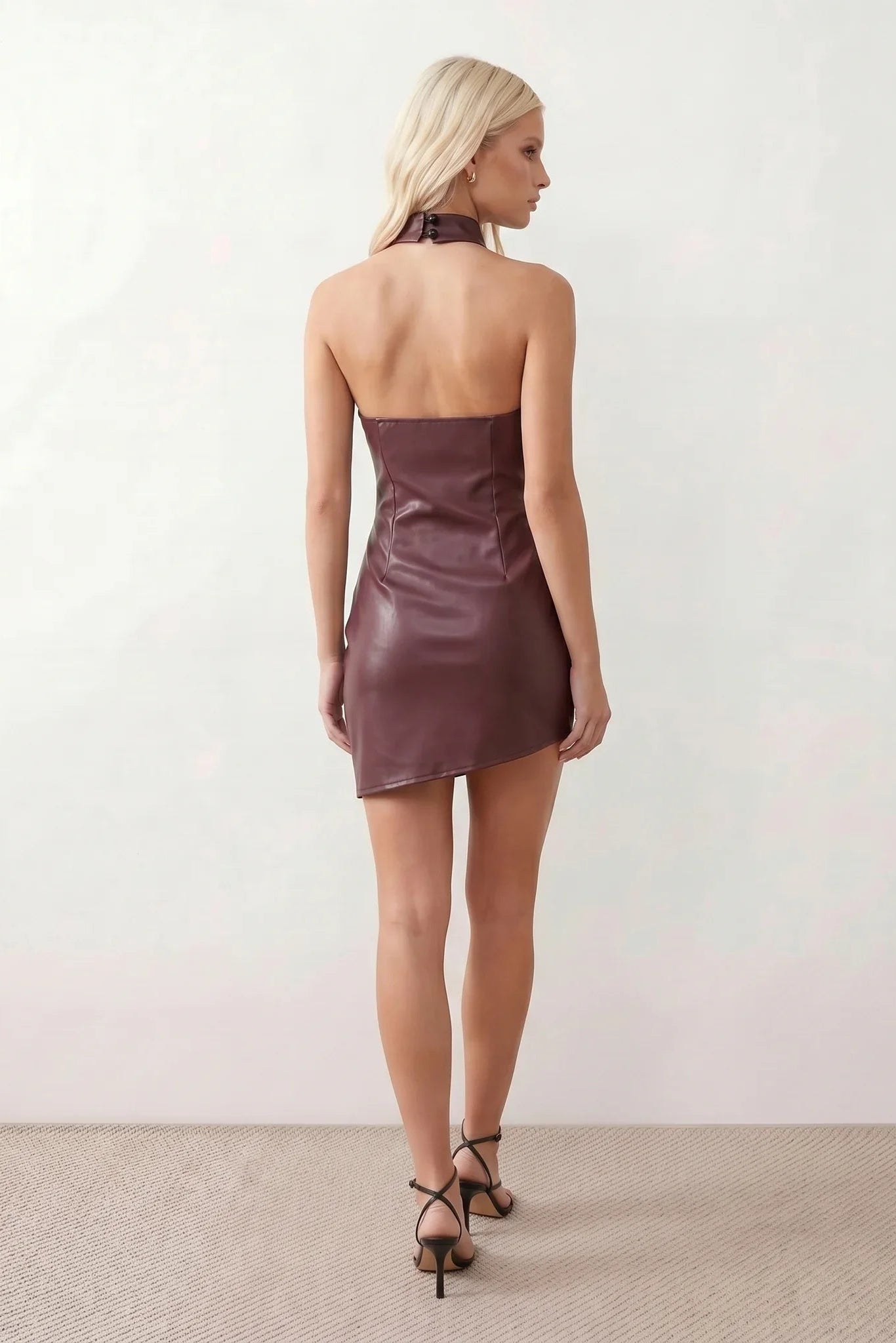 Woman wearing a glossy faux leather halter mini dress with a draped cowl neckline, fully backless design, ruched waist and hips, bodycon fit, and an asymmetric mini hem, styled for parties, cocktail events, and evening occasions.