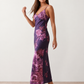 Woman wearing the Milota glossy floral slip maxi dress featuring a baroque-inspired floral print, deep V-neckline, adjustable spaghetti straps, low draped back, satin-look finish, and body-skimming floor-length silhouette, ideal for wedding guests and formal occasions.