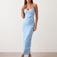 Woman wearing the Milota glossy floral slip maxi dress featuring a baroque-inspired floral print, deep V-neckline, adjustable spaghetti straps, low draped back, satin-look finish, and body-skimming floor-length silhouette, ideal for wedding guests and formal occasions.