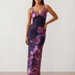 Woman wearing the Milota glossy floral slip maxi dress featuring a baroque-inspired floral print, deep V-neckline, adjustable spaghetti straps, low draped back, satin-look finish, and body-skimming floor-length silhouette, ideal for wedding guests and formal occasions.