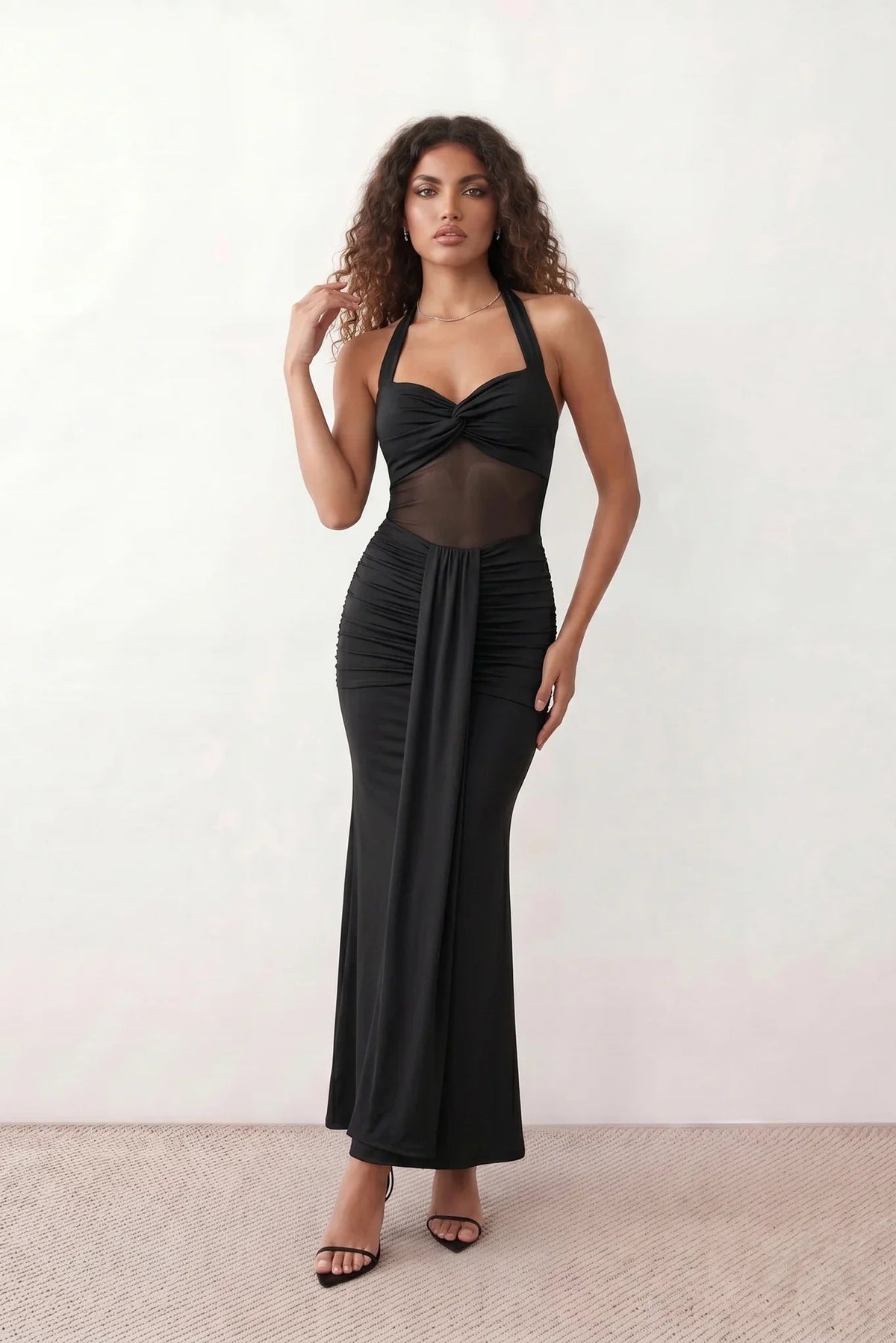 Woman wearing the Halter Neck Mermaid Maxi Dress featuring a sweetheart twist-knot neckline, sheer mesh waist panel, ruched mermaid silhouette, draped front skirt, open back with halter tie fastening, and elegant floor-length finish.