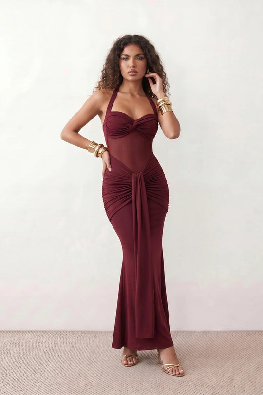 Woman wearing the Halter Neck Mermaid Maxi Dress featuring a sweetheart twist-knot neckline, sheer mesh waist panel, ruched mermaid silhouette, draped front skirt, open back with halter tie fastening, and elegant floor-length finish.