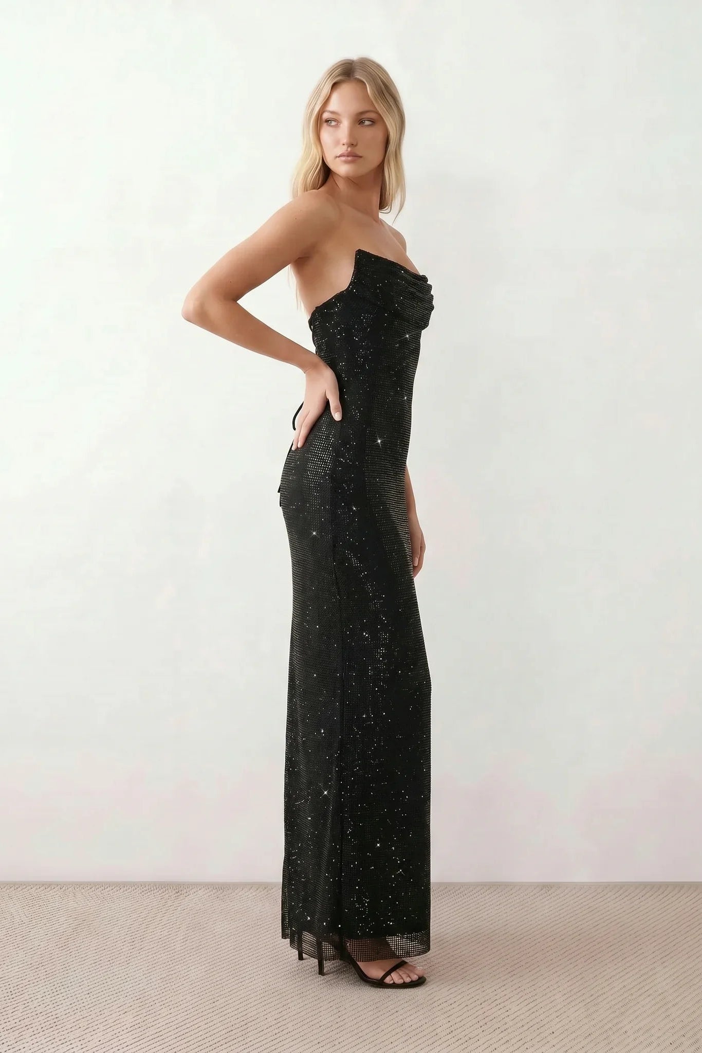 Woman wearing the Izabela sequined strapless maxi dress featuring shimmering all-over sequins, sparkling rhinestone embellishments, a ruched cowl-fold bust, adjustable lace-up corset back and a figure-skimming column silhouette with a floor-length hem, perfect for formal evening occasions.