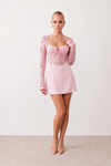 Woman wearing the Joaquina Lace Sweetheart Mini Dress in delicate lace-look fabric with a sweetheart neckline, long sleeves, an open lace-up back with bow detail, a defined waist and a flattering mini-length silhouette.