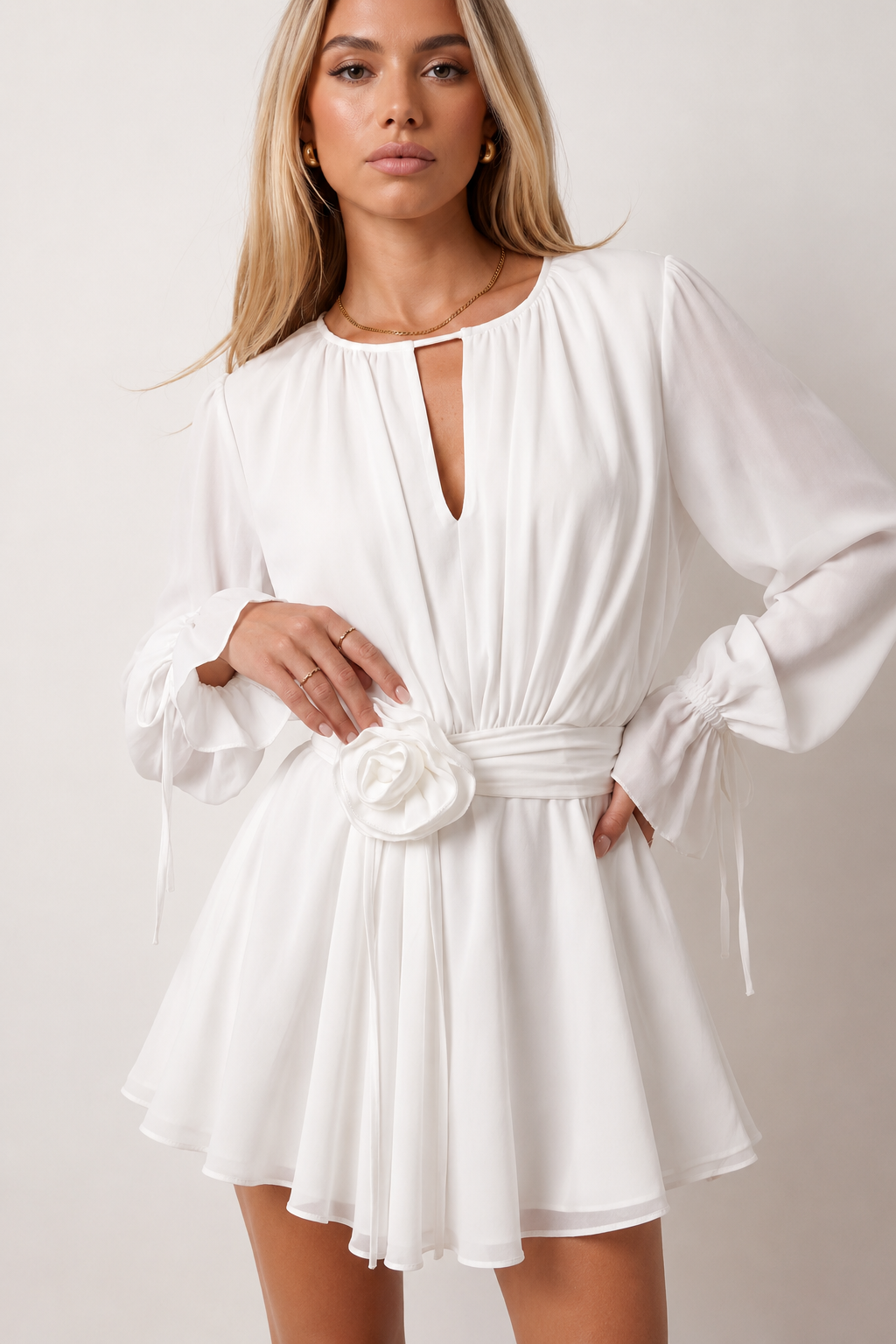 Woman wearing the Michela pleated tie detail mini dress featuring lightweight pleated fabric, a keyhole neckline with ribbon tie, billowy long sleeves with ribbon cuffs, a 3D floral corsage at the waist, belted sash, open low back and a double-layer ruffled hem, ideal for weddings, garden parties and special occasions.