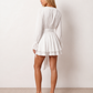 Woman wearing the Michela pleated tie detail mini dress featuring lightweight pleated fabric, a keyhole neckline with ribbon tie, billowy long sleeves with ribbon cuffs, a 3D floral corsage at the waist, belted sash, open low back and a double-layer ruffled hem, ideal for weddings, garden parties and special occasions.