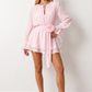 Woman wearing the Michela pleated tie detail mini dress featuring lightweight pleated fabric, a keyhole neckline with ribbon tie, billowy long sleeves with ribbon cuffs, a 3D floral corsage at the waist, belted sash, open low back and a double-layer ruffled hem, ideal for weddings, garden parties and special occasions.