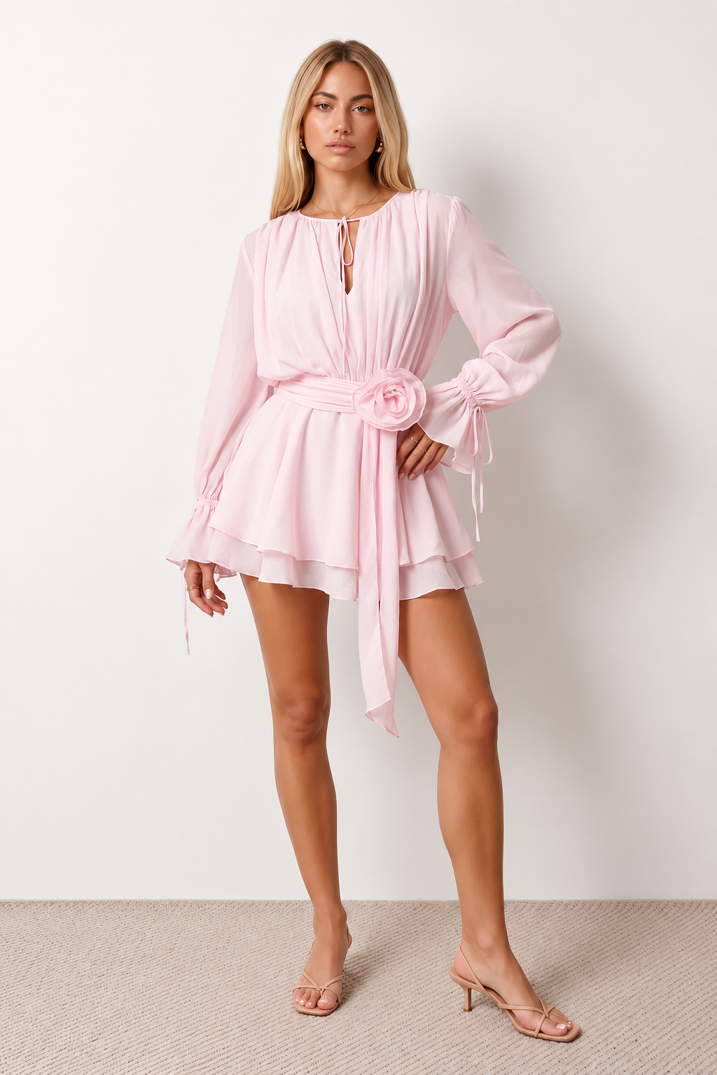 Woman wearing the Michela pleated tie detail mini dress featuring lightweight pleated fabric, a keyhole neckline with ribbon tie, billowy long sleeves with ribbon cuffs, a 3D floral corsage at the waist, belted sash, open low back and a double-layer ruffled hem, ideal for weddings, garden parties and special occasions.
