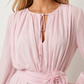 Woman wearing the Michela pleated tie detail mini dress featuring lightweight pleated fabric, a keyhole neckline with ribbon tie, billowy long sleeves with ribbon cuffs, a 3D floral corsage at the waist, belted sash, open low back and a double-layer ruffled hem, ideal for weddings, garden parties and special occasions.