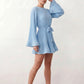 Woman wearing the Pleated Ruffle Detail Mini Dress featuring an all-over pleated design, flowing bell sleeves, a round neckline, elasticated waist with a self-tie sash, and a feminine ruffle hem, perfect for weddings, garden parties, race days, and elegant occasions.