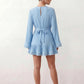 Woman wearing the Pleated Ruffle Detail Mini Dress featuring an all-over pleated design, flowing bell sleeves, a round neckline, elasticated waist with a self-tie sash, and a feminine ruffle hem, perfect for weddings, garden parties, race days, and elegant occasions.