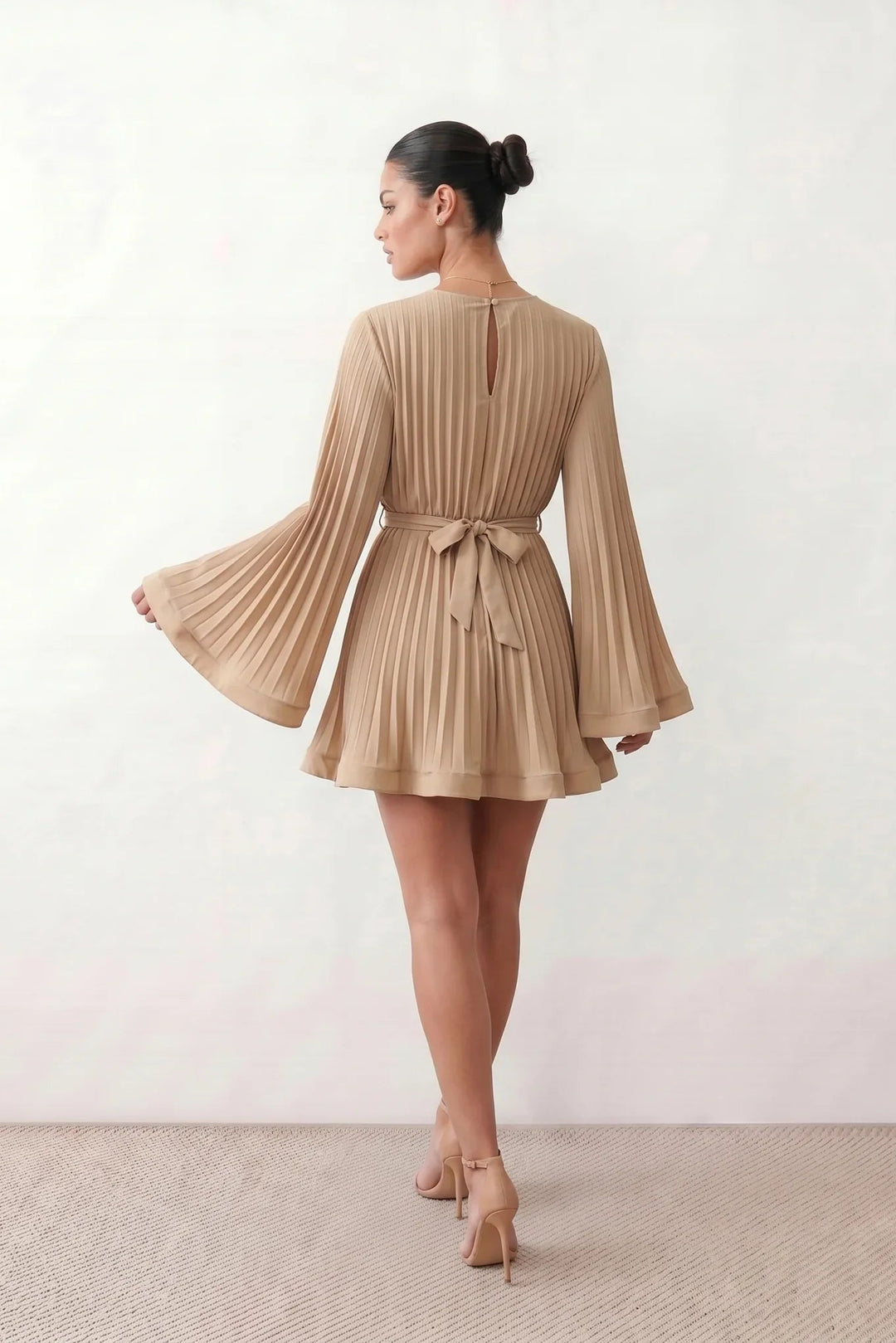 Woman wearing the Pleated Ruffle Detail Mini Dress featuring an all-over pleated design, flowing bell sleeves, a round neckline, elasticated waist with a self-tie sash, and a feminine ruffle hem, perfect for weddings, garden parties, race days, and elegant occasions.