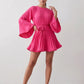 Woman wearing the Pleated Ruffle Detail Mini Dress featuring an all-over pleated design, flowing bell sleeves, a round neckline, elasticated waist with a self-tie sash, and a feminine ruffle hem, perfect for weddings, garden parties, race days, and elegant occasions.