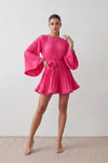Woman wearing the Pleated Ruffle Detail Mini Dress featuring an all-over pleated design, flowing bell sleeves, a round neckline, elasticated waist with a self-tie sash, and a feminine ruffle hem, perfect for weddings, garden parties, race days, and elegant occasions.