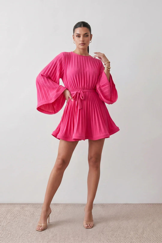 Woman wearing the Pleated Ruffle Detail Mini Dress featuring an all-over pleated design, flowing bell sleeves, a round neckline, elasticated waist with a self-tie sash, and a feminine ruffle hem, perfect for weddings, garden parties, race days, and elegant occasions.